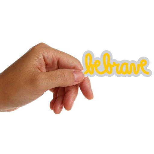 Hand holding a 'be brave' sticker by Matthew Hoffman with yellow text on a white background