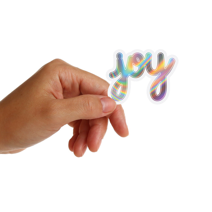 Hand holding a holographic 'joy' sticker by Matthew Hoffman against a white background