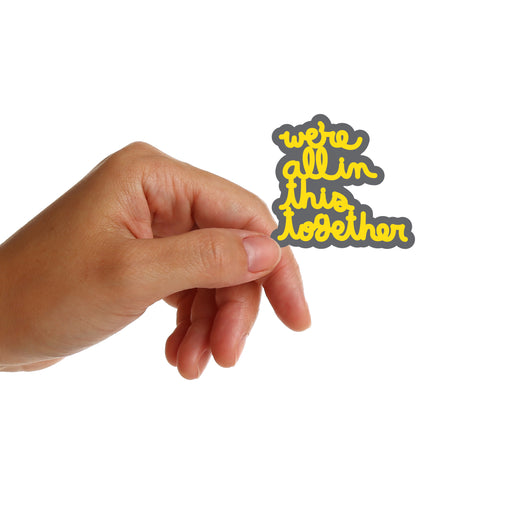 Hand holding a sticker with 'we're all in this together' sticker by Matthew Hoffman on a white background
