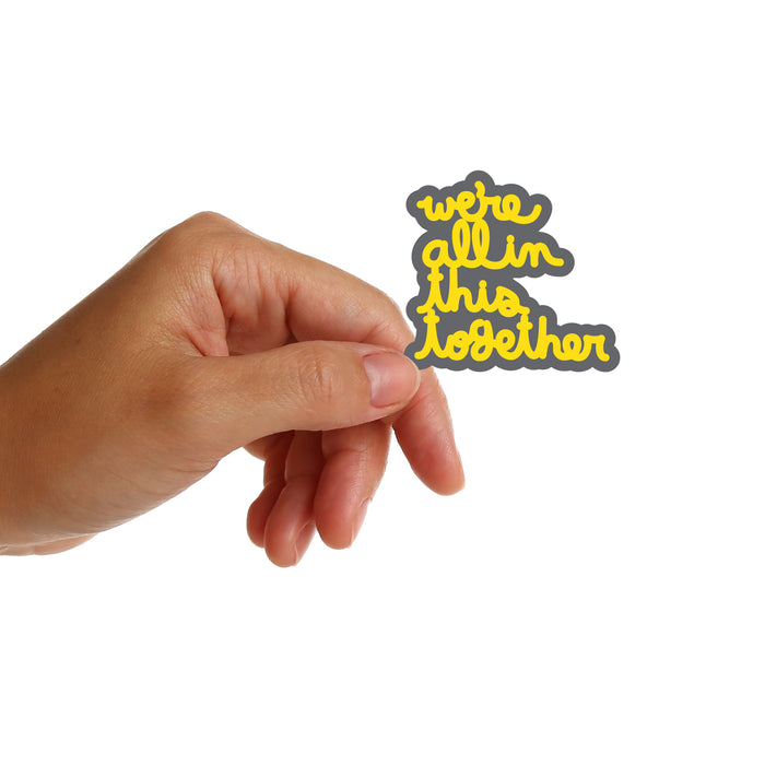 Hand holding a sticker with 'we're all in this together' sticker by Matthew Hoffman on a white background