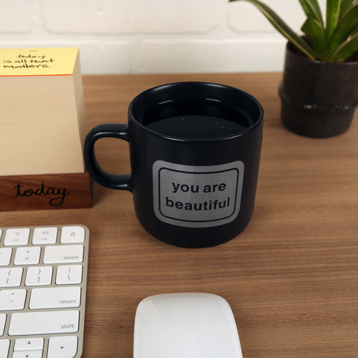 Matte black ceramic mug with the classic You Are Beautiful sticker icon on one side, designed by Matthew Hoffman.