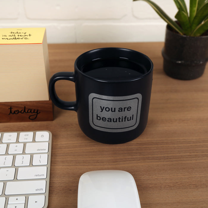 Matte black ceramic mug with the classic You Are Beautiful sticker icon on one side, designed by Matthew Hoffman.